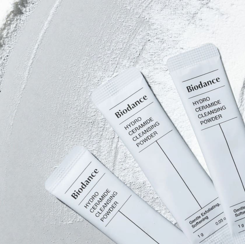 Три саше засобу BIODANCE Hydro Ceramide Cleansing Powder лежать на поверхні, на якій розсипаний порошок.