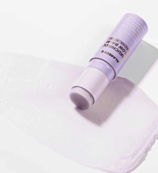 Відкритий рожевий стік LALARECIPE Bakuchinol Glow Balm лежить на білій поверхні, поруч — сліди його текстури.