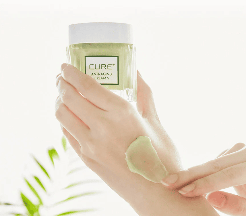Рука тримає баночку крему Cure Anti-Aging Cream пальцями, на шкірі видно розмазану кремову текстуру зеленуватого кольору.