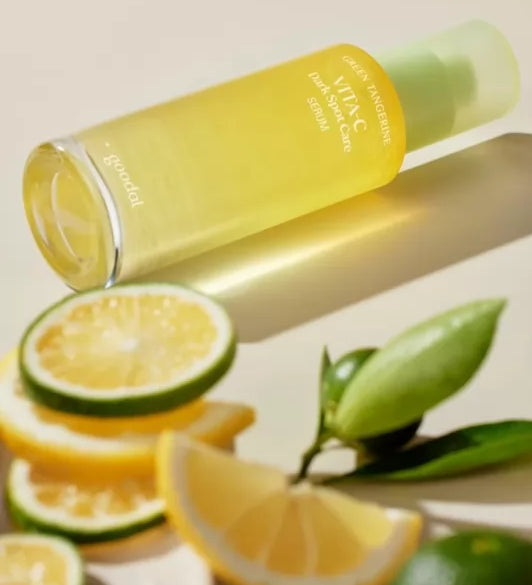 Пляшечка Goodal Green Tangerine Vita C Dark Spot Care Serum лежить на білій поверхні, поруч з нею видно шматочки лимону та лайму.
