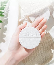 Твердий шампунь LABO-H у круглій формі бежевого кольору з темними вкрапленнями, лежить на долоні