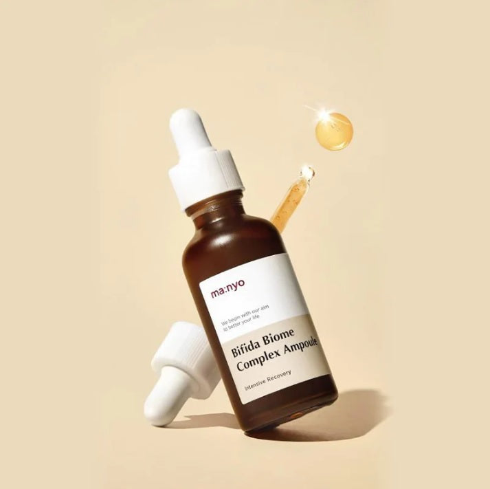 Пляшечка Ma:nyo Bifida Biome Essence зависла у повітрі, поряд перевернута піпетка, з якої капає есенція на бежевому фоні.