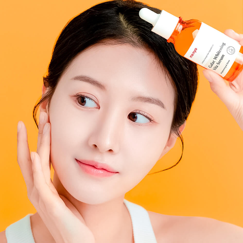 Дівчина азіатської зовнішності тримає ампулу Ma:nyo Galac Whitening Vita Serum біля голови, з легкою посмішкою на обличчі.