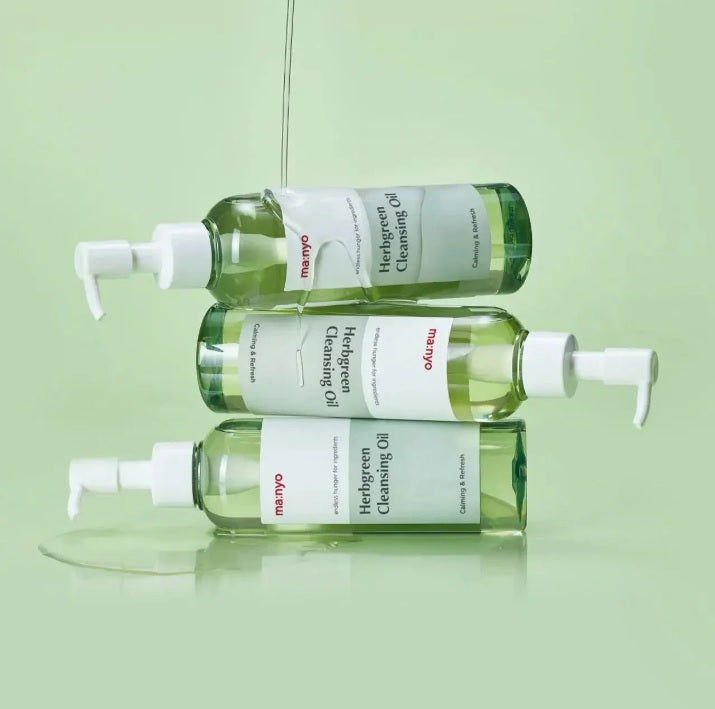 На зеленій поверхні одна на одній лежать пляшки засобу Ma:nyo Herb Cleansing Oil, на них зверху розливається його текстура.