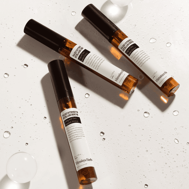 Три ампули сироватки NARD Long And Glam Intensive Eyelash Serum лежать на білій поверхні, вкритій крапельками води.