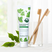 На білій поверхні стоїть тюбик зубної пасти NARD Whitening Toothpaste, поряд склянка і дві дерев'яні щітки для зубів, на фоні видно рослину.