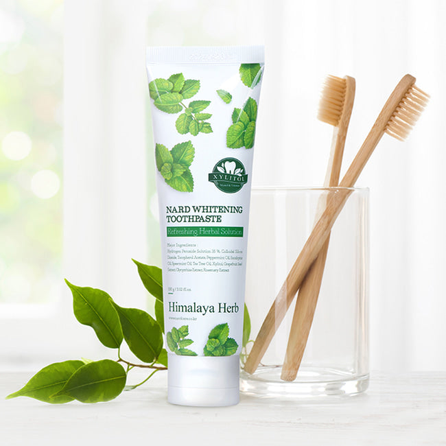 На білій поверхні стоїть тюбик зубної пасти NARD Whitening Toothpaste, поряд склянка і дві дерев'яні щітки для зубів, на фоні видно рослину.