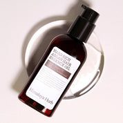 Темно-прозорий флакон з чорним дозатором олії-есенції для волосся Nard Rich Argan Hair Essence Oil лежить на скляній тарілочці на білій поверхні