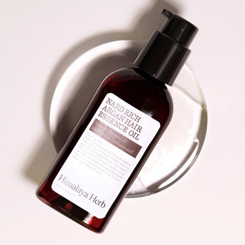 Темно-прозорий флакон з чорним дозатором олії-есенції для волосся Nard Rich Argan Hair Essence Oil лежить на скляній тарілочці на білій поверхні