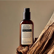 Темно-прозорий флакон з чорним дозатором олії-есенції для волосся Nard Rich Argan Hair Essence Oil стоїть на частині кори дерева.