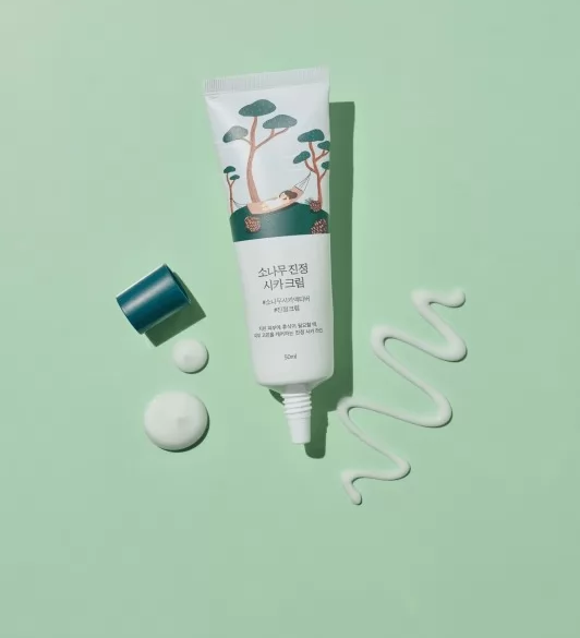 Відкритий тюбик крему Round Lab Pine Calming Cica Cream на зеленій поверхні, поряд видно нанесену текстуру крему, а також кришечку тюбика.