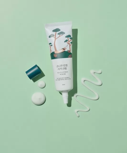 Відкритий тюбик крему Round Lab Pine Calming Cica Cream на зеленій поверхні, поряд видно нанесену текстуру крему, а також кришечку тюбика.