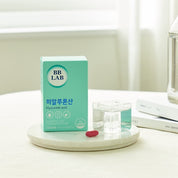 На білому подіумі стоїть коробка з колагеном BB Lab Hyaluronic Acid та червона капсула, на фоні книги та пляшки.