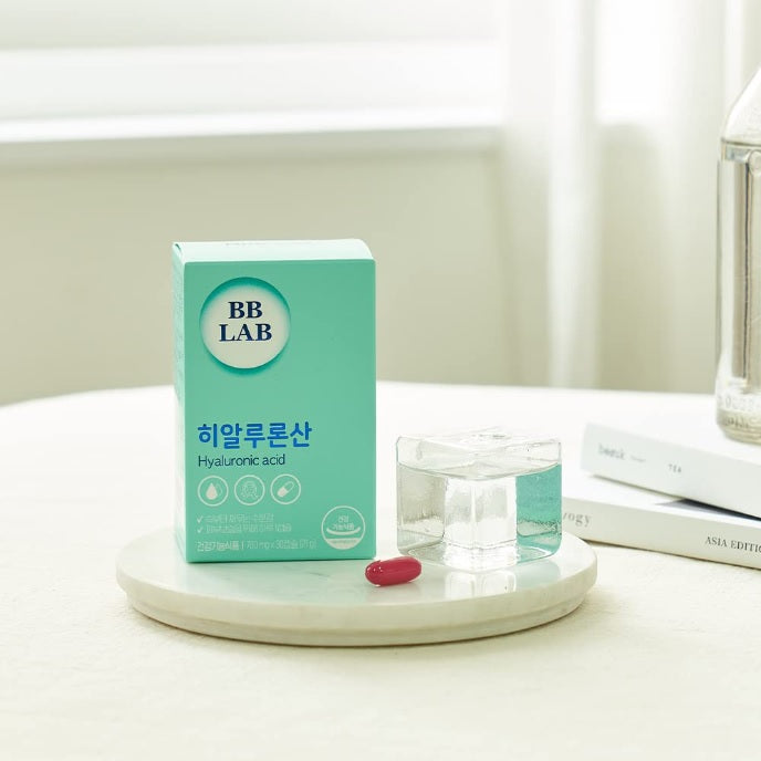 На білому подіумі стоїть коробка з колагеном BB Lab Hyaluronic Acid та червона капсула, на фоні книги та пляшки.
