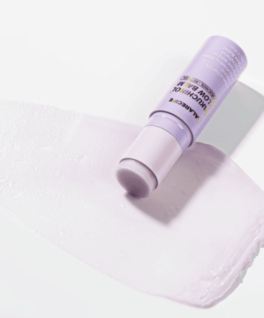Відкритий рожевий стік LALARECIPE Bakuchinol Glow Balm лежить на білій поверхні, поруч — сліди його текстури.