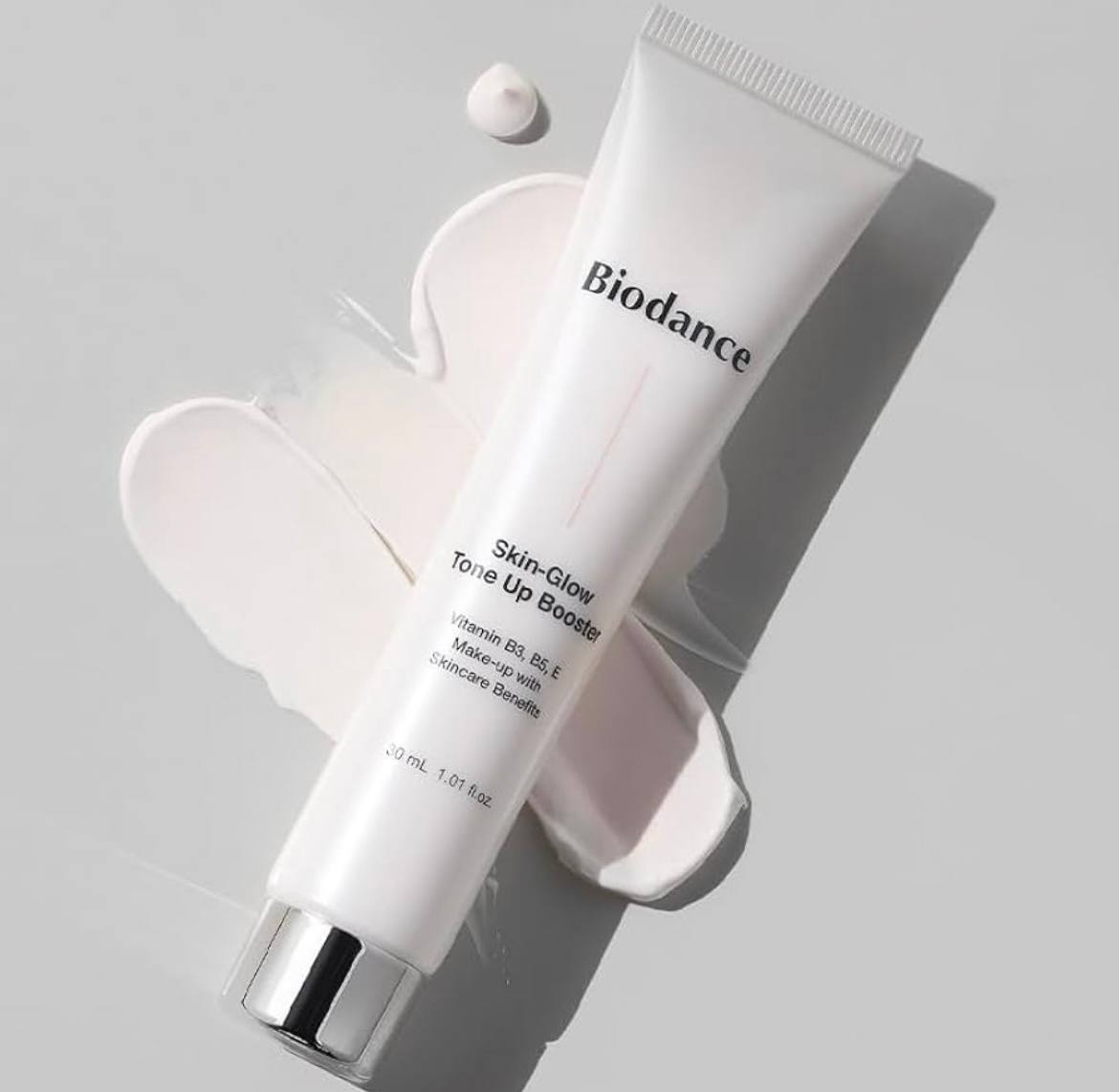 Білий тюбик Biodance Skin Glow Tone Up Booster 30 мл з чорними написами та сріблястою кришечкою лежить на кремовій текстурі, нанесеній на білу поверхню