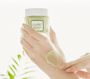 Рука тримає баночку крему Cure Anti-Aging Cream пальцями, на шкірі видно розмазану кремову текстуру зеленуватого кольору.