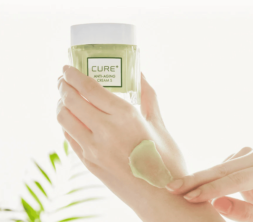 Рука тримає баночку крему Cure Anti-Aging Cream пальцями, на шкірі видно розмазану кремову текстуру зеленуватого кольору.