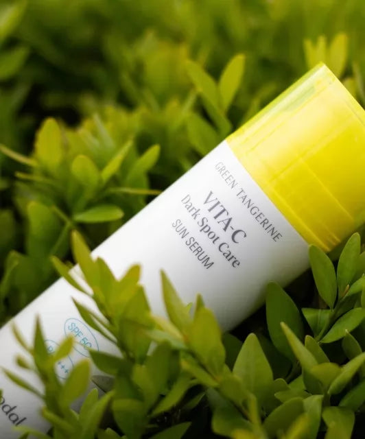 Біла пляшка з жовтою кришкою Goodal Green Tangerine Vita C Dark Spot Sun Care Serum SPF 50+ PA++++ лежить на зеленій рослинності.