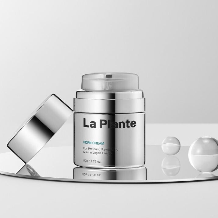 Відкрита срібляста баночка крему LA PLANTE PDRN Cream 50 g стоїть на скляній поверхні, поруч — дві прозорі кульки, наполовину заповнені рідиною.