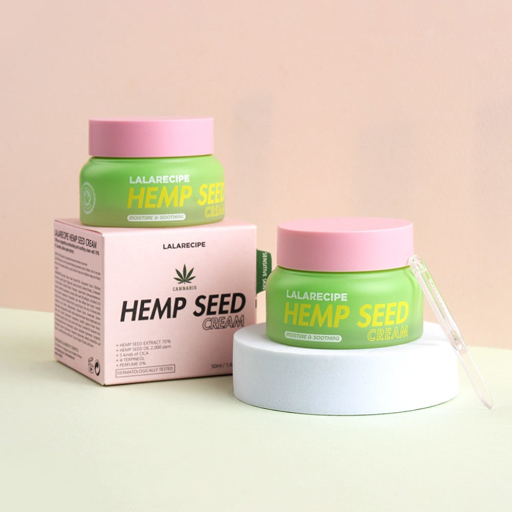 Дві зелені баночки з рожевою кришкою крему LALARECIPE Hemp Seed Cream: одна стоїть на білій платформі, інша — на коробці від крему.