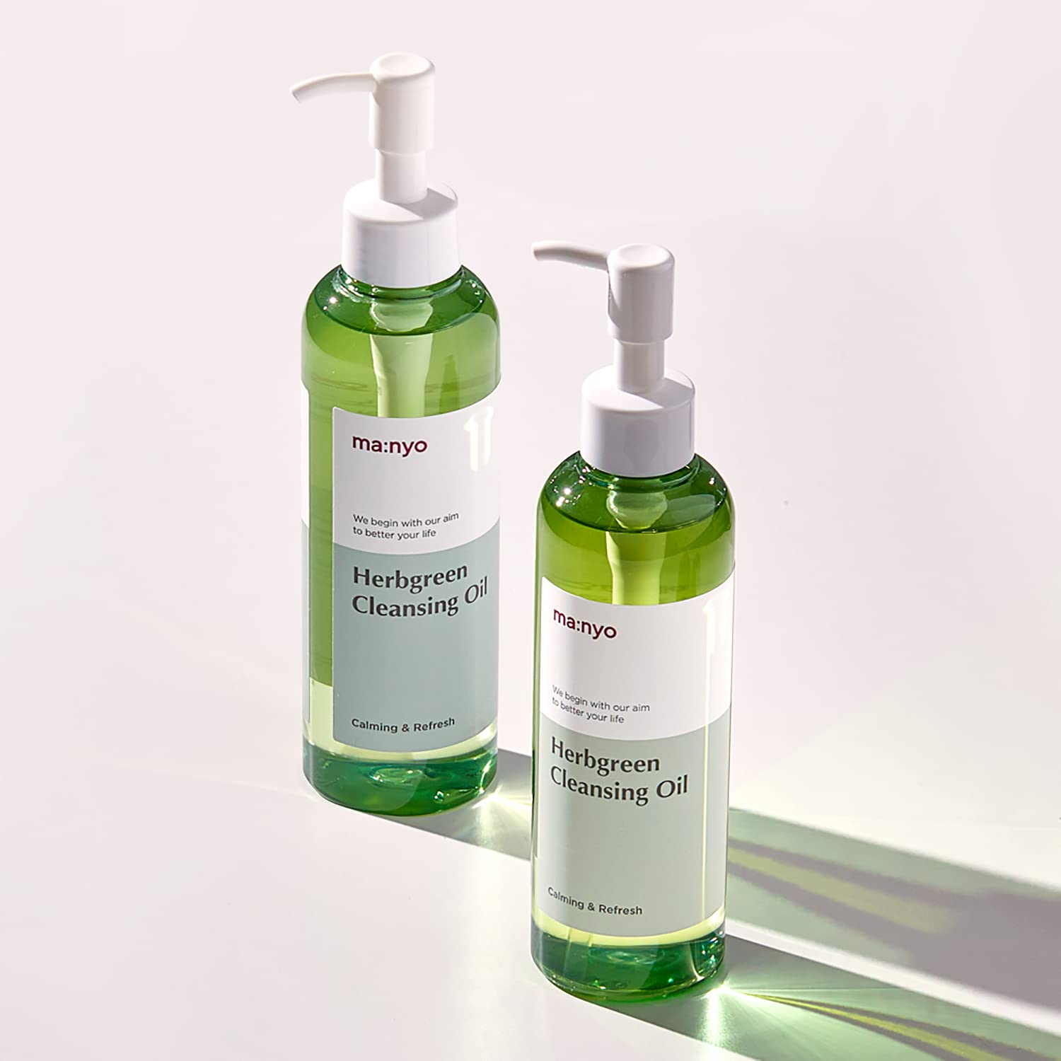 Дві пляшечки засобу Ma:nyo Herb Cleansing Oil стоять на білий поверхні, на них світить яскраве світло.