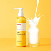 Жовта пляшечка засобу Ma:nyo Pure Cleansing Milk з білим дозатором стоїть на жовтій поверхні та жовтому фоні, поряд склянка, в яку наливається молоко.