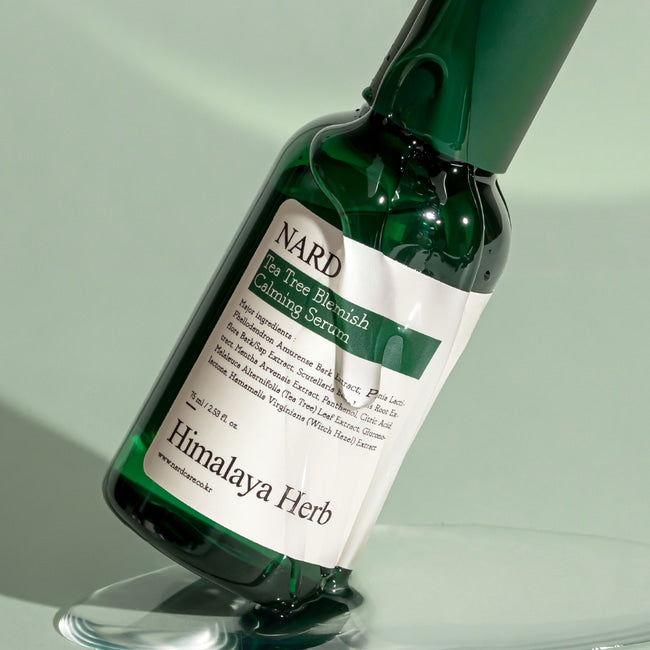 Ампула Bouquet Garni Nard Tea Tree Blemish Calming Serum стоїть під кутом на білій поверхні та вкрита есенцією, яка розтеклася навколо.