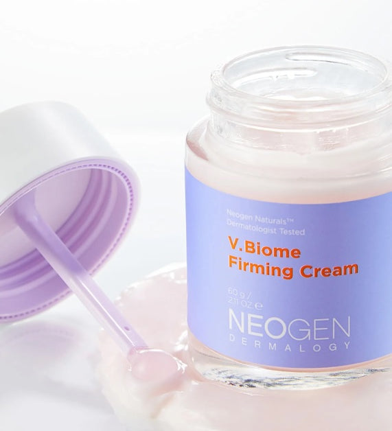 Баночка крему Neogen Dermalogy V.Biome Firming Cream стоїть на поверхні, покритій ніжним шаром крему, поруч лежить кришечка.