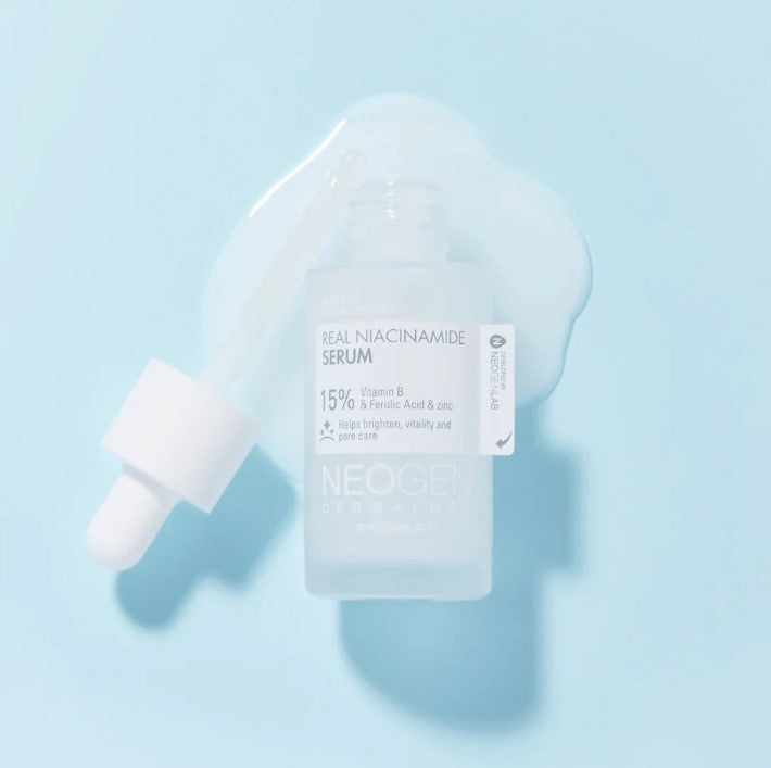 Ампула сироватки Neogen Dermalogy Real Niacinamide 15% Firming Serum лежить відкрита поряд з піпеткою в розлитій текстурі на блакитній поверхні.