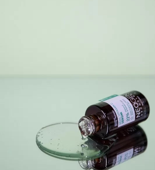 Ампула сироватки Neogen Dermalogy Real Vitamin лежить на дзеркальній поверхні, з відкритого горлечка витікає прозора сироватка.