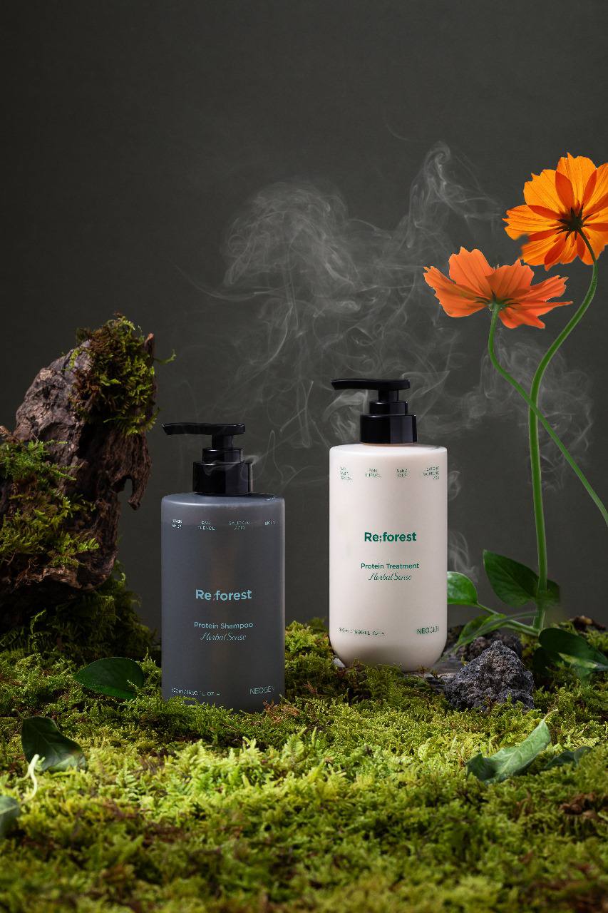 Сірий шампунь та білий кондиціонер Neogen Re;forest з чорними дозаторами на траві серед каменів з оранжевими квітками та легким димом.
