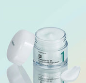 Відкрита скляна баночка з білою кришкою крему Numbuzin No.1 Pantothenic B5 Active Soothing Cream стоїть біля розмазаної текстури крему, кришка оперта на баночку.