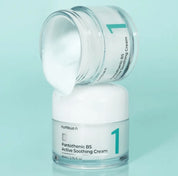Дві скляні баночки крему Numbuzin No.1 Pantothenic B5 Active Soothing Cream: одна стоїть, інша лежить на першій, відкрита, з якої витікає крем, на бірюзовому фоні.