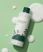 Альт текст: Світло-зелена баночка лосьйону ROUND LAB Pine Calming Cica Lotion лежить на зеленій поверхні, поруч розлита, білими плямами, текстура лосьйону