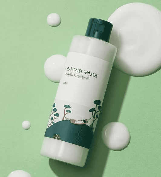 Альт текст: Світло-зелена баночка лосьйону ROUND LAB Pine Calming Cica Lotion лежить на зеленій поверхні, поруч розлита, білими плямами, текстура лосьйону