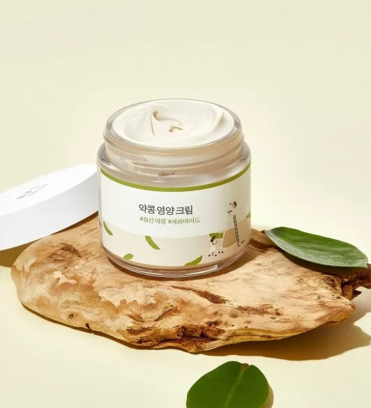 Відкрита баночка крему Round Lab Soybean Nourishing Cream стоїть на камені, поруч з нею видно пару зелених листочків.