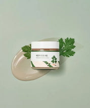 Баночка крему Round Lab Mugwort Calming Cream лежить на кремі та зеленій рослині на ярко-зеленій поверхні.