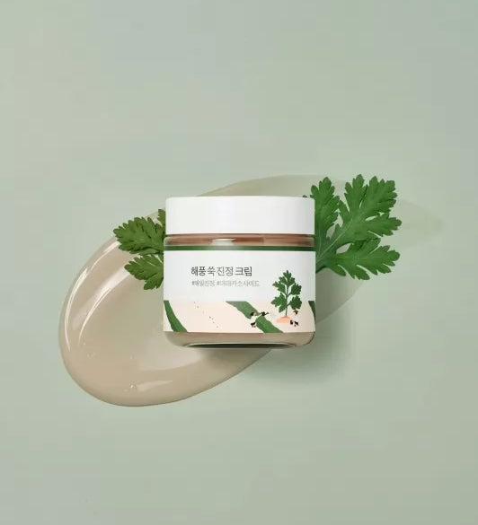 Баночка крему Round Lab Mugwort Calming Cream лежить на кремі та зеленій рослині на ярко-зеленій поверхні.