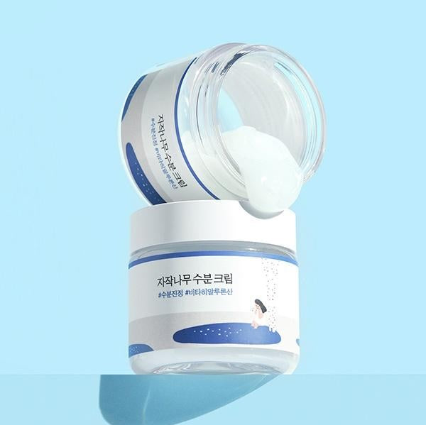 Дві баночки крему Round Lab Juice Moisturizing Cream: одна стоїть, інша — відкрита, лежить зверху, і з неї витікає крем на блакитному фоні.