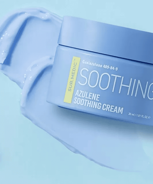 На фото баночка крему Azulene Soothing Cream лежить на текстурі крему, яка знаходиться на синій поверхні.