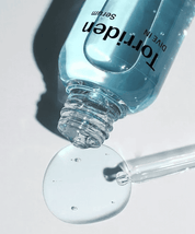 Відкрита ампула Torriden Dive-In Low Molecular Hyaluronic Acid Skin Serum лежить на білій поверхні, з неї витікає блакитна сироватка, поруч лежить піпетка.