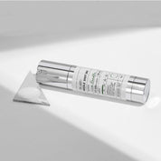 Бустер VT Cosmetics VT Reedle Shot 300 лежить на білій поверхні, поруч — трикутне дзеркальце з нанесеною текстурою засобу.